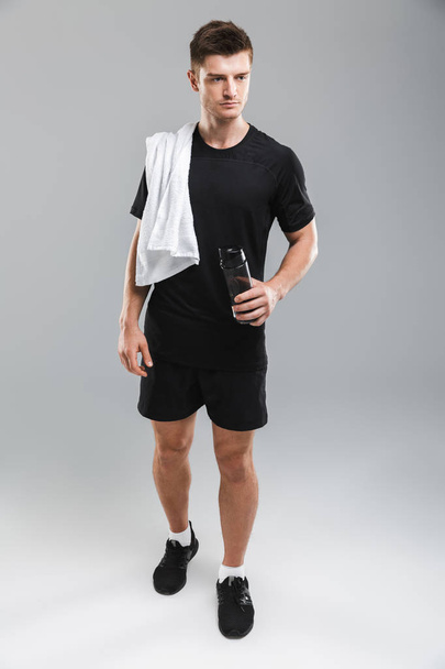 Full length portrait of a serious young sportsman standing with water bottle and towel on his shoulder isolated over gray background - Foto, immagini