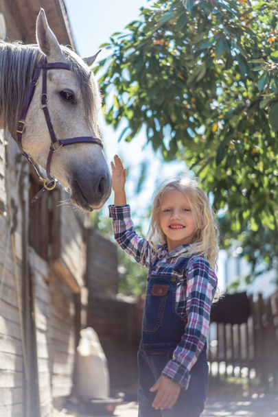 smiling kid touching white horse at farm and looking at camera - Photo, Image