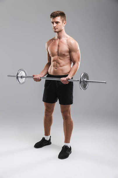 Full length portrait of a concentrated shirtless sportsman doing exercises with a barbell isolated over gray background - Foto, Imagem