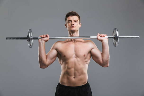 Portrait of a strong shirtless sportsman doing exercises with a barbell isolated over gray background - Foto, imagen