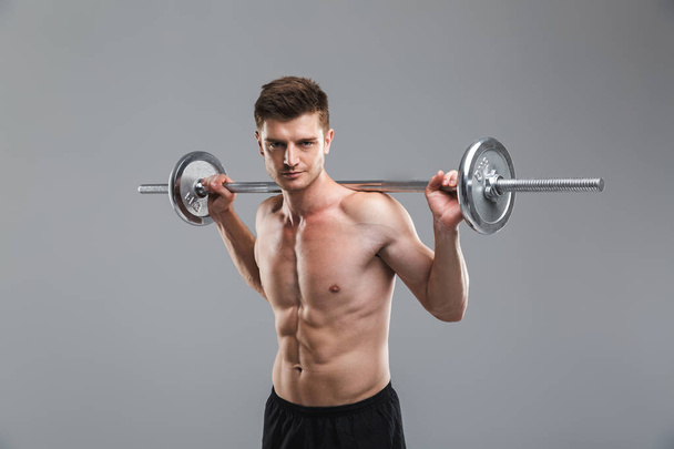 Portrait of a confident shirtless sportsman doing exercises with a barbell isolated over gray background - Foto, Bild
