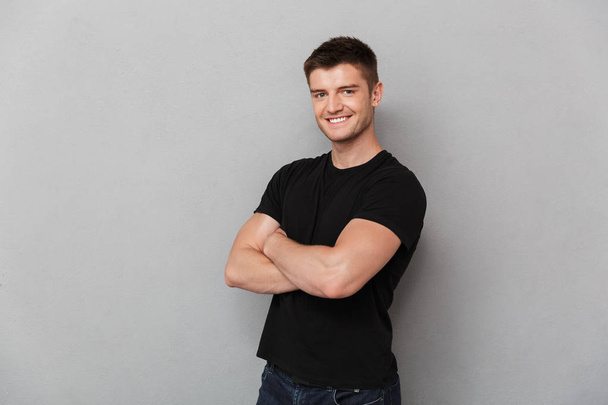 Portrait of a smiling young man standing with arms folded isolated over gray background - Photo, Image