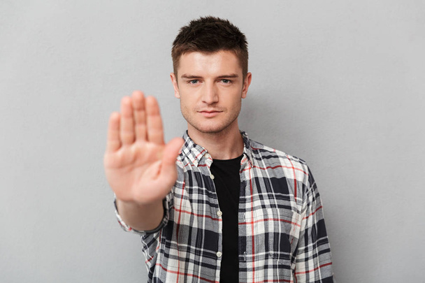 Portret van een ernstige jonge man tonen stop gebaar met zijn palm over grijze achtergrond - Foto, afbeelding
