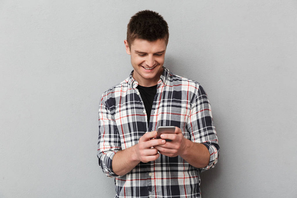 Portrait of a smiling young man using mobile phone over gray background - Φωτογραφία, εικόνα