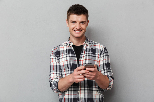 Portrait of a smiling young man holding mobile phone over gray background - Fotografie, Obrázek