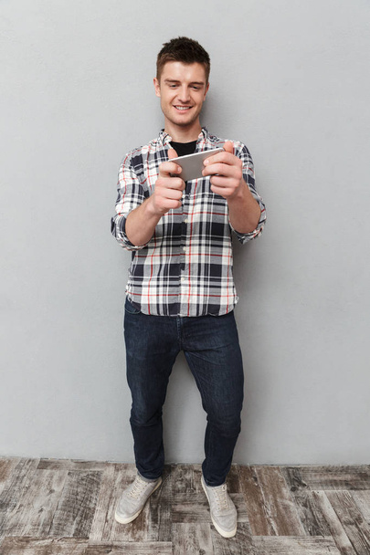 Ολόσωμο πορτρέτο του ένας χαμογελαστός νεαρός παίζει παιχνίδια στο κινητό πάνω από το γκρίζο φόντο - Φωτογραφία, εικόνα