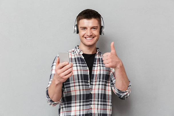 Portrait of a satisfied young man listening to music with headphones and holding mobile phone over gray background - Фото, изображение