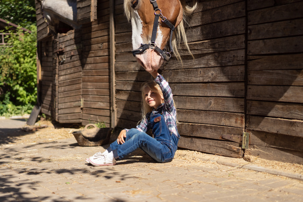 dziecko siedzi na ziemi w pobliżu stajni i dotykając konia w gospodarstwie - Zdjęcie, obraz