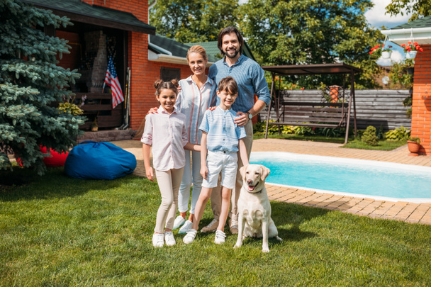 χαρούμενη οικογένεια με σκυλί λαμπραντόρ βλέπουν φωτογραφική μηχανή, ενώ στέκεται στην πίσω αυλή του σπιτιού χώρας θερινή ημέρα - Φωτογραφία, εικόνα
