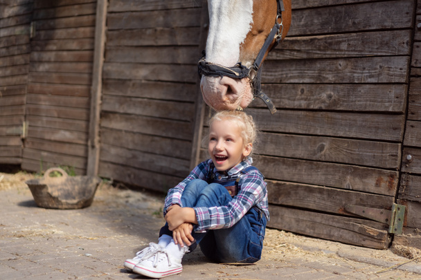 happy kid sitting on ground and horse touching her hair at farm - Foto, Bild