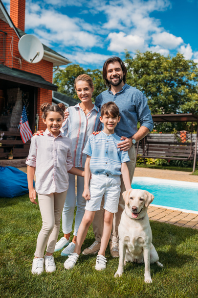 familia alegre con perro labrador mirando a la cámara mientras está de pie en el patio trasero de la casa de campo en el día de verano
 - Foto, imagen