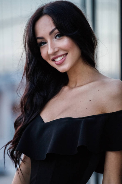 Portrait of beautiful young brunette woman in black dress with stylish makeup standing outdoors at daytime, blurred street background - Foto, immagini