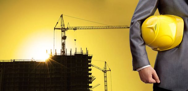 inżynier, trzymając żółty kask dla bezpieczeństwa pracowników na tle nowego budynku i budowa żurawi na tle wieczorem zachód słońca niebo pochmurne - Zdjęcie, obraz