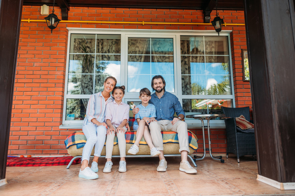 uśmiechnięta rodzina siedzi na kanapie razem na ganku domu w kraju  - Zdjęcie, obraz