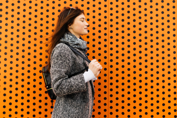 lovely dark-haired city girl in a gray trendy coat posing against the background of an unusual orange wall - Zdjęcie, obraz