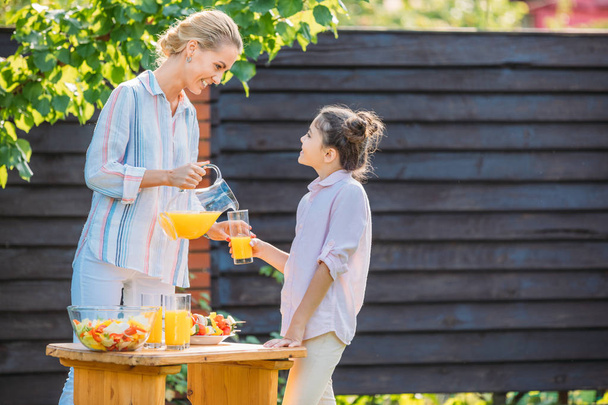 side view of smiling mother pouring juice into daughters glass during picnic on backyard - Photo, Image