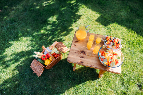 top view of basket with food for picnic, glasses of  juice and fresh salad on wooden chair on green lawn - Photo, Image