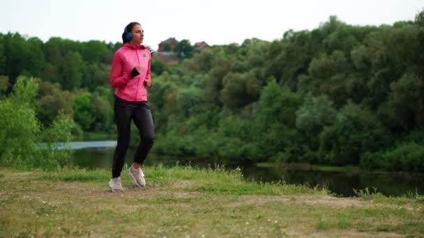 Uma menina de casaco rosa e calças pretas corre perto do rio em fones de ouvido se preparando para a maratona
. - Filmagem, Vídeo