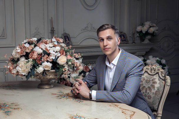 Dear young man with a haircut in a suit, sitting at a table in a room with a classic interior. Luxury. Male beauty, fashion. Vintage. Composition from flowers. - Photo, Image