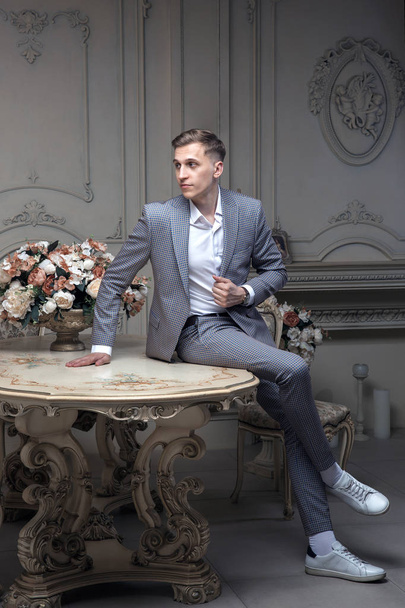Dear young man with a haircut in a suit, sitting at a table in a room with a classic interior. Luxury. Male beauty, fashion. Vintage. Composition from flowers. - Fotó, kép