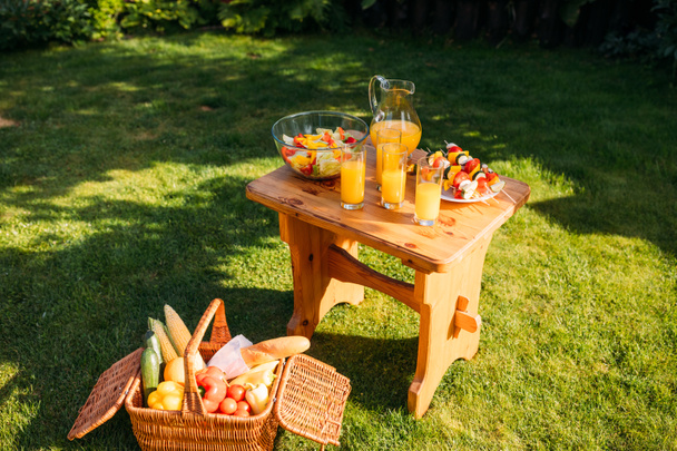 Nahaufnahme von Korb mit Picknick-Essen, Gläsern Saft und frischem Salat auf Holzstuhl auf grünem Rasen - Foto, Bild