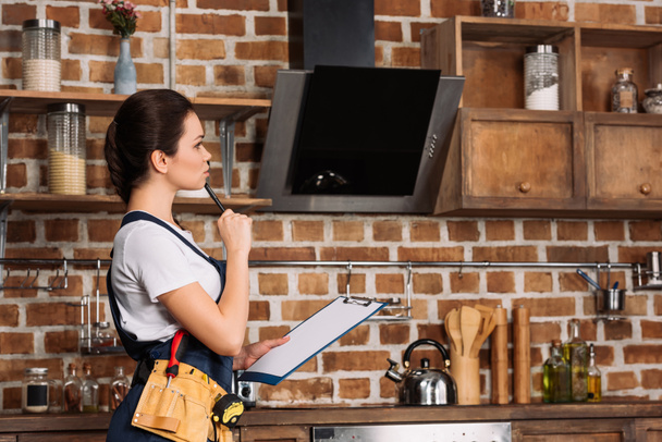 thoughtful young repairwoman with clipboard standing at kitchen and looking away - Photo, Image