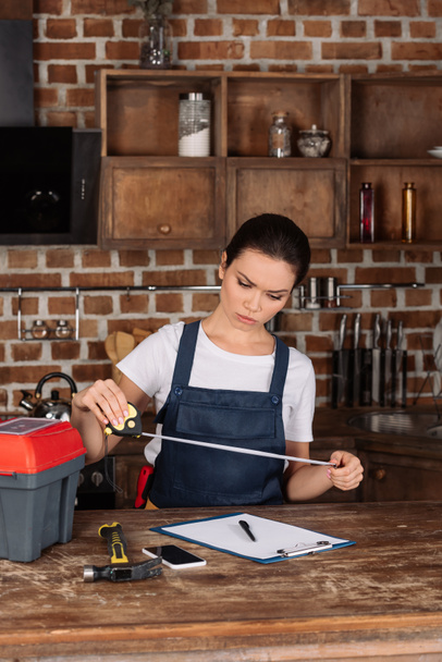 serious young repairwoman with measuring tape and clipboard at kitchen - Photo, Image