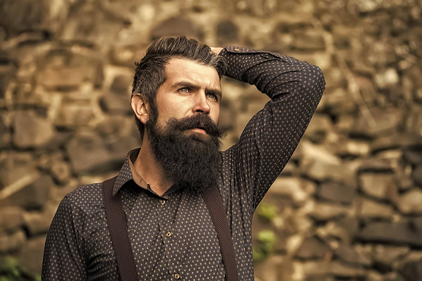 Male revelation. Closeup view of one handsome senior man with black hair and long lush beard in blue shirt and purple trausers standing outdoor on stone wall grey background, horizontal picture - Valokuva, kuva