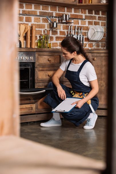 serious young repairwoman with clipboard sitting near oven and looking at it - Foto, Imagem