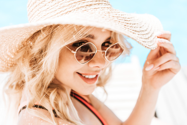 portrait of beautiful young blonde woman in wicker hat and sunglasses smiling at camera near pool   - Photo, Image