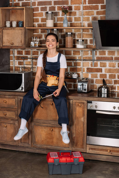 glückliche junge Reparaturfrau in Uniform sitzt am Küchentisch und blickt in die Kamera - Foto, Bild