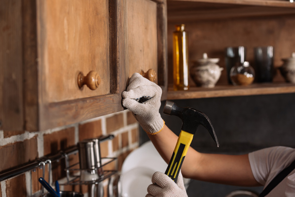 cropped shot of repairwoman in work gloves hammering nail into kitchen cabinet - 写真・画像