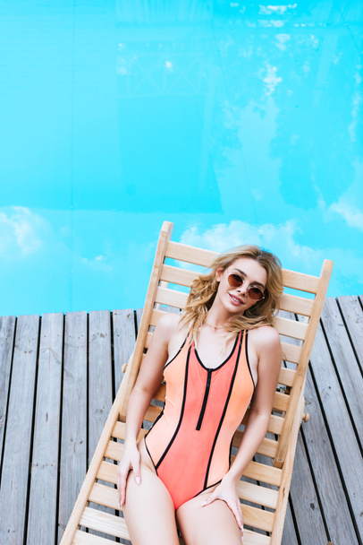 high angle view of beautiful blonde woman in swimsuit and sunglasses resting on chaise lounge near pool   - Fotoğraf, Görsel