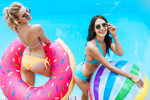 high angle view of happy young women in bikini and sunglasses holding inflatable ball and doughnut in swimming pool   - Fotografie, Obrázek