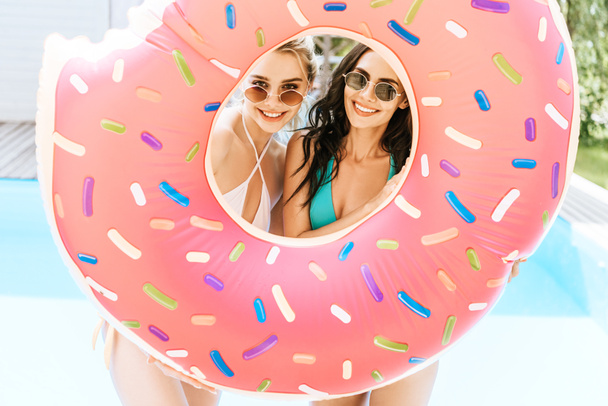 jeunes femmes heureuses en lunettes de soleil et maillots de bain tenant un beignet de bain et souriant à la caméra près de la piscine
 - Photo, image