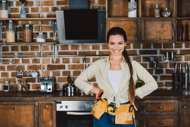 glücklich junge Reparaturfrau mit Armen akimbo steht in der Küche und blickt in die Kamera - Foto, Bild