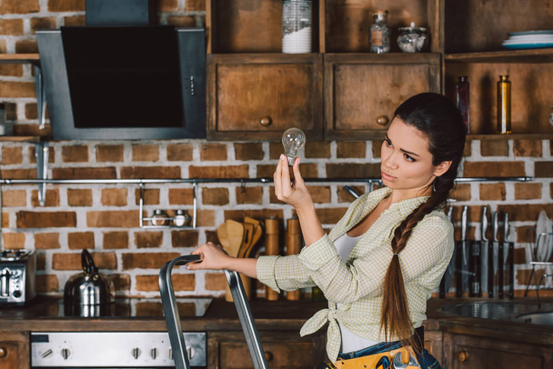 confident young repairwoman standing on stepladder and examining light bulb at kitchen - Photo, Image