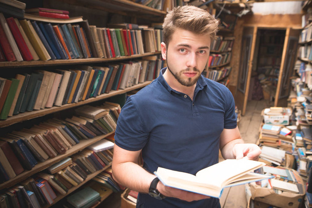 Porträt eines Mannes, der mit einem Buch in der Hand in einer öffentlichen Bibliothek steht und in die Kamera blickt. ein Student wählt Literatur in einer alten öffentlichen Bibliothek aus. - Foto, Bild