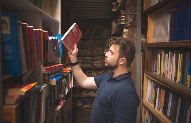 Portrait of a man with a beard who takes books from a shelf in a library. Student chooses books in an old public library. Search for books in an atmospheric library. - Photo, Image