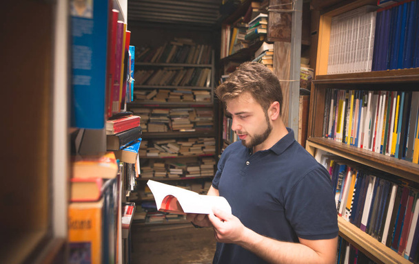 Szép szakállas ember áll egy hangulatos nyilvános könyvtár a keze és olvas egy könyvet. Egy diák keres egy könyvet a könyvtárban. A választás az irodalom régi nyilvános könyvtár. - Fotó, kép