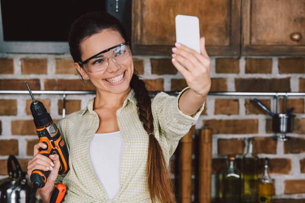 Glückliche junge Reparaturfrau in Schutzbrille mit Bohrmaschine macht Selfie - Foto, Bild