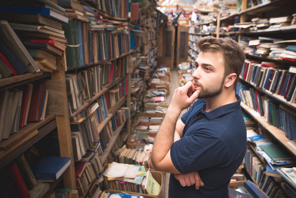 Retrato de un hombre con barba parado en el fondo de una biblioteca pública atmosférica y mirando la estantería. El estudiante selecciona libros en una biblioteca. Búsqueda de literatura en una biblioteca pública
 - Foto, Imagen
