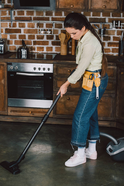 atractiva mujer joven con cinturón de herramientas utilizando aspiradora en la cocina
 - Foto, imagen