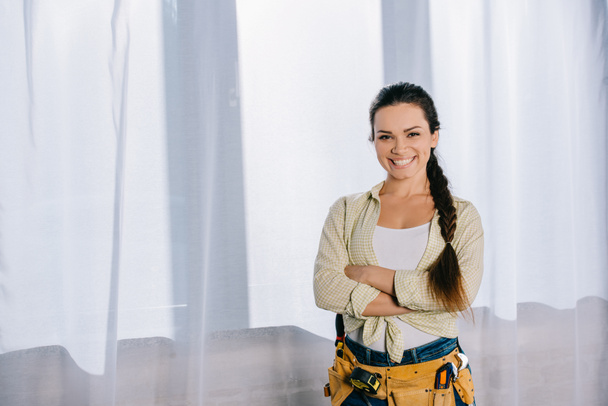 beautiful young repairwoman with toolbelt and crossed arms looking at camera - Photo, Image