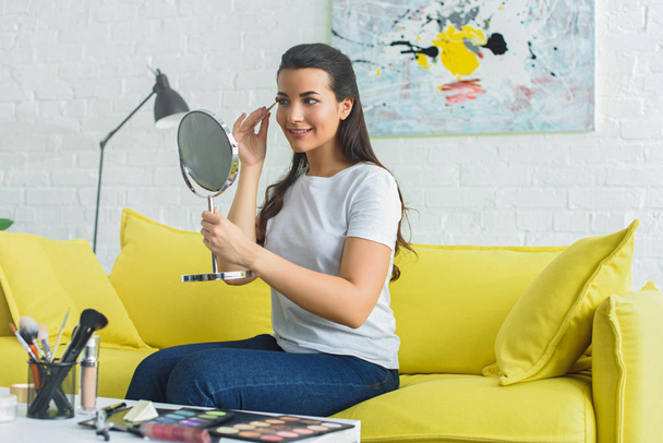 νεαρός χαμογελαστό γυναίκα κοιτάζοντας καθρέφτη και εφαρμόζοντας σκιές ματιών ενώ κάνει μακιγιάζ στον καναπέ στο σπίτι - Φωτογραφία, εικόνα