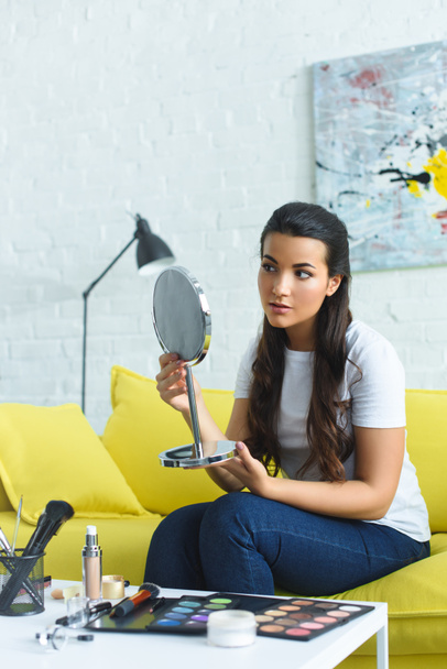 young beautiful woman looking at mirror while sitting on sofa at coffee table with cosmetics at home - Foto, Imagen