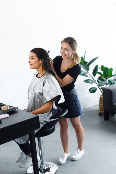 side view of smiling hairstylist with comb doing hairstyle for young woman  - Photo, image