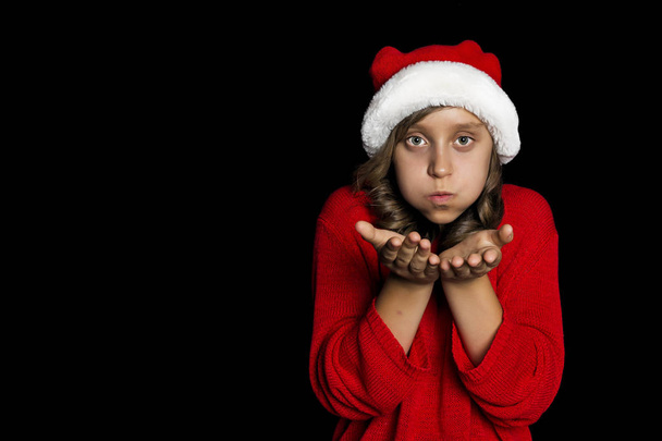 A young girl in a red sweater and a Santa Claus hat blows on her hands and looks at the viewer. Black isolate. Copy space. - Фото, зображення