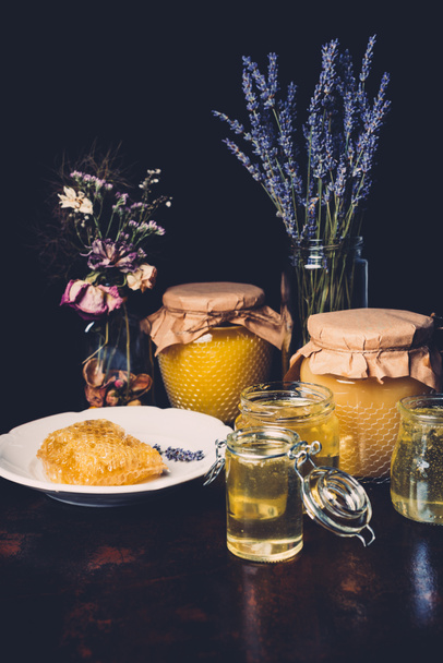 selective focus of jars with honey, dried flowers, lavender and honeycomb on black background  - Foto, immagini