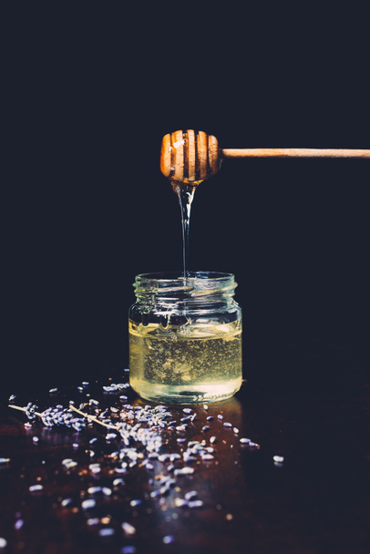 selective focus of honey stick with flowing honey into jar surrounded by lavender petals on black background  - Photo, Image